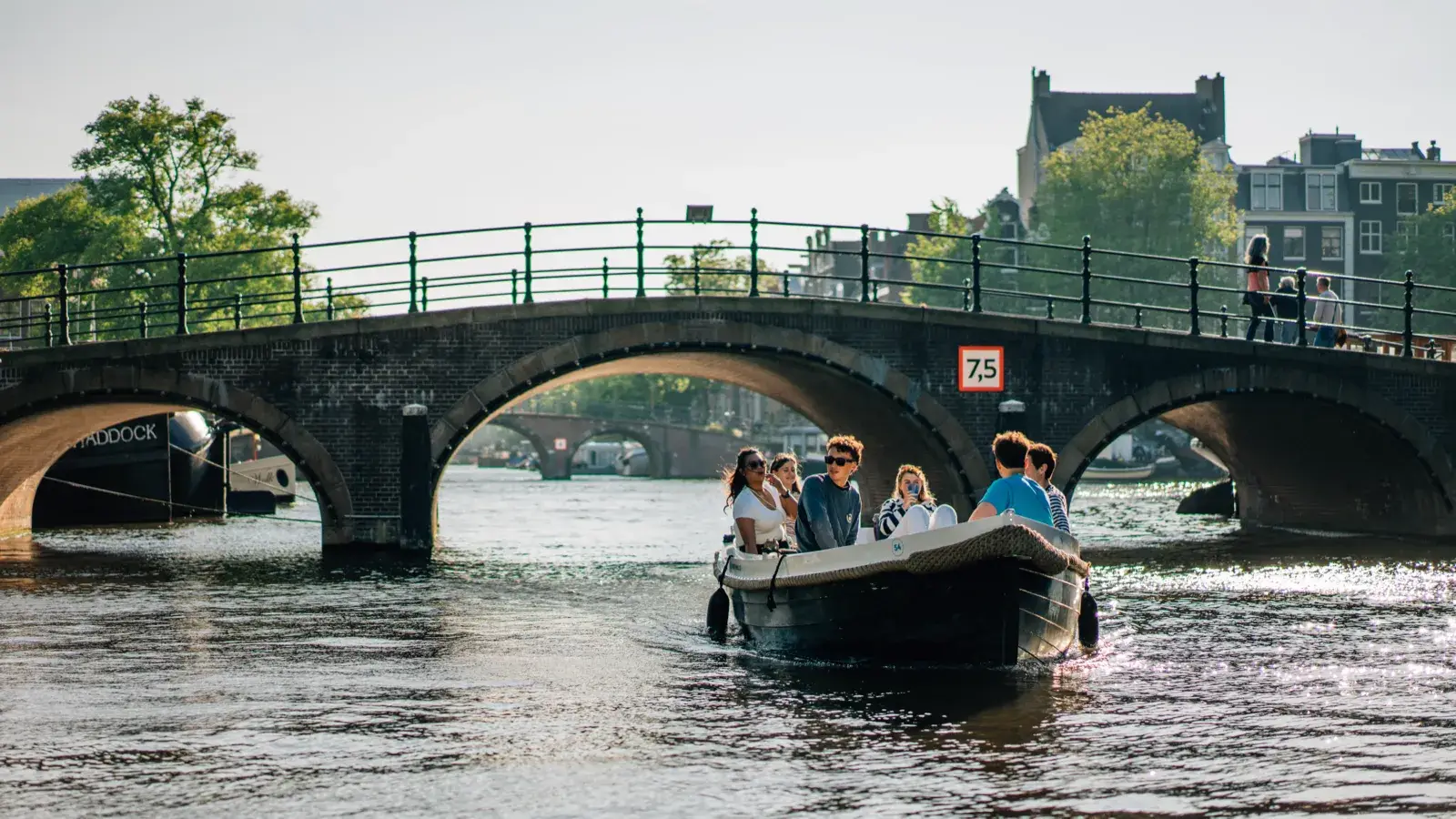 Friends in an open rental motorboat on an Amsterdam canal, passing under a stone bridge