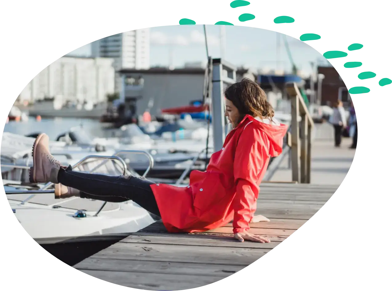 A woman in a red raincoat sits on a wooden dock with her feet up on the bow of a moored motorboat