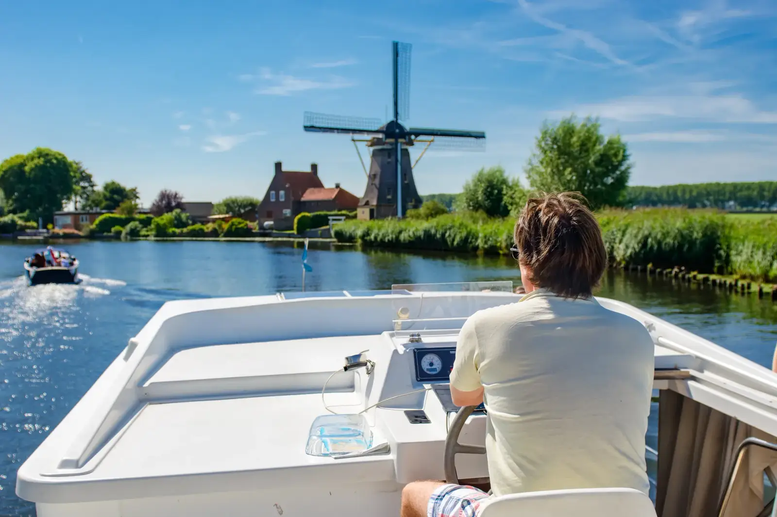 Ein Mann steuert ein weißes Hausboot über einen breiten niederländischen Fluss, vorbei an einer Windmühle und einem Bauernhof