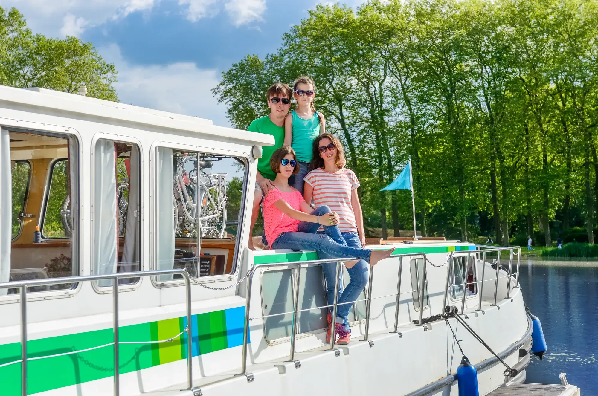 Eine Familie an Deck eines Hausboots am Ufer, die Fahrräder in der Kajüte verstaut