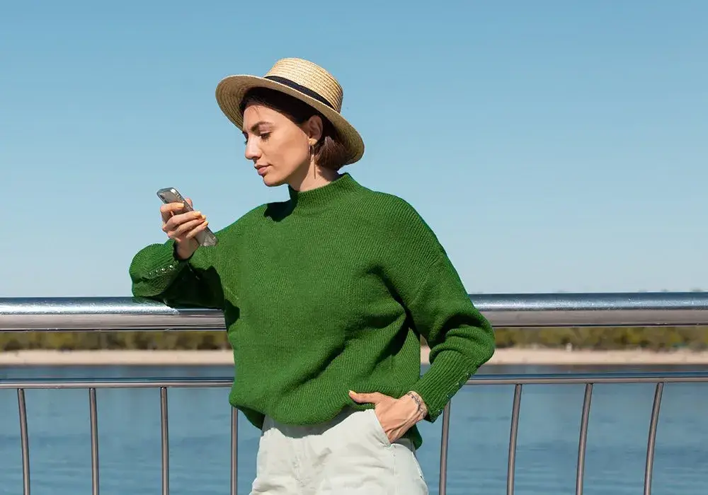 A woman in a green sweater and a straw hat checks her phone at a railing by the water