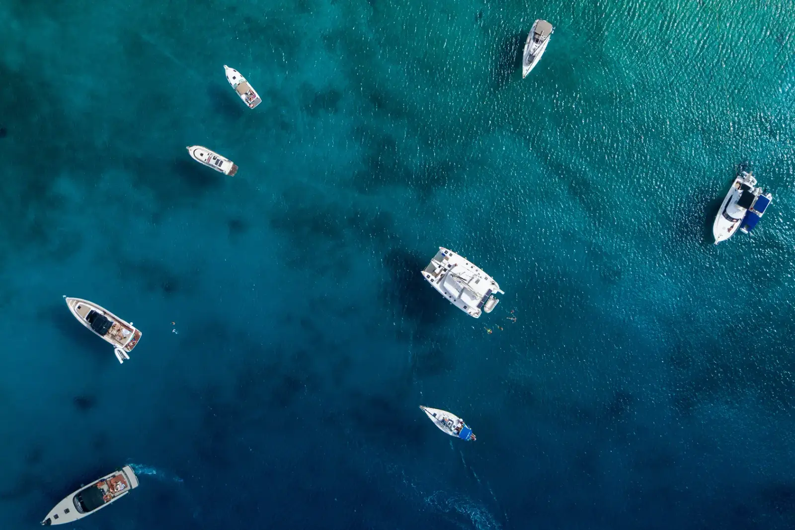Des bateaux au mouillage dans une eau turquoise et claire, vus d'en haut, avec un catamaran au milieu et des yachts à moteur autour