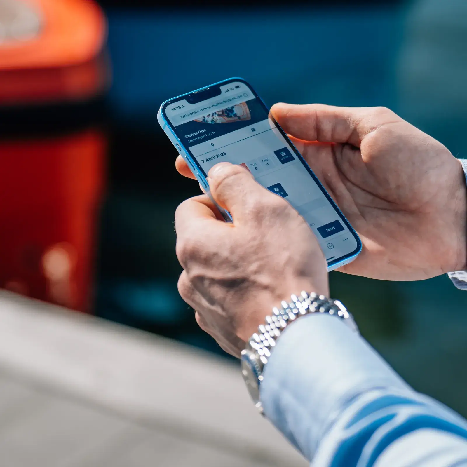 Hands holding a phone with a boat booking open, beside a red boat at the dock