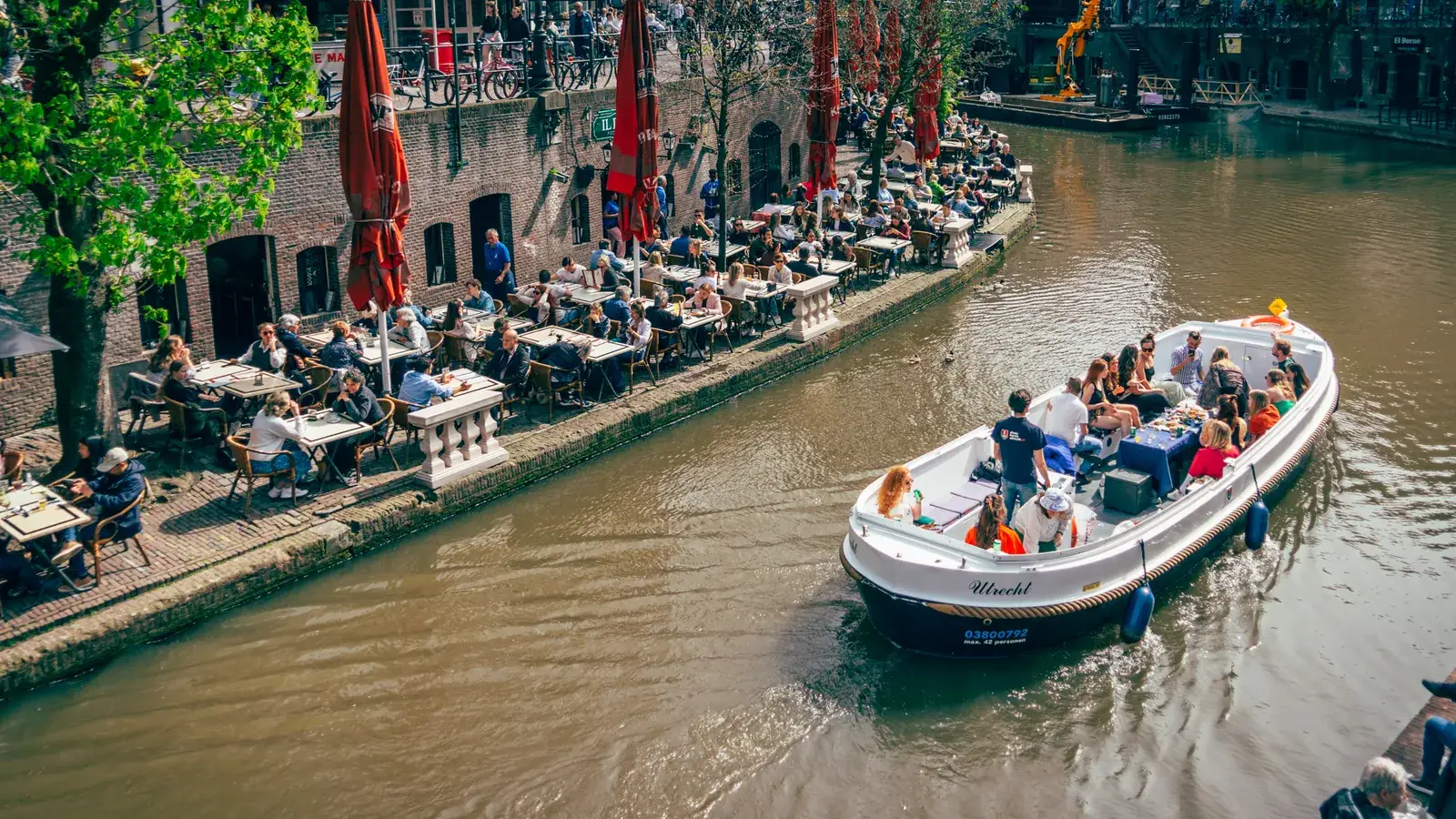 An open canal boat full of guests passes the wharf terraces in Utrecht