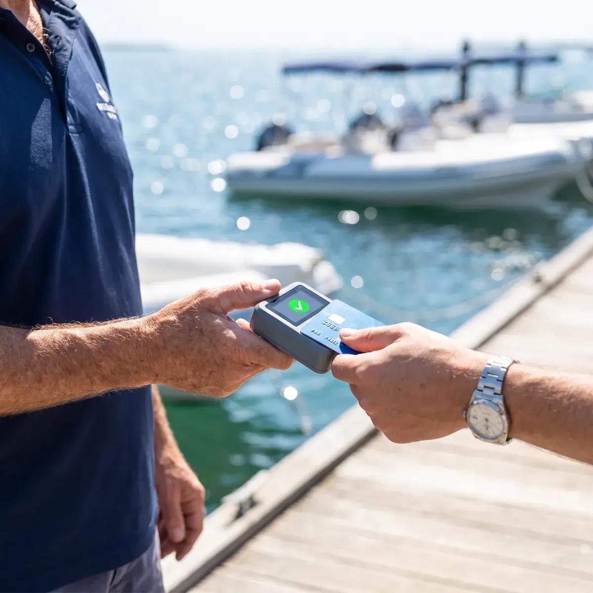 On a sunny jetty, a staff member holds out a card reader and a customer taps to pay