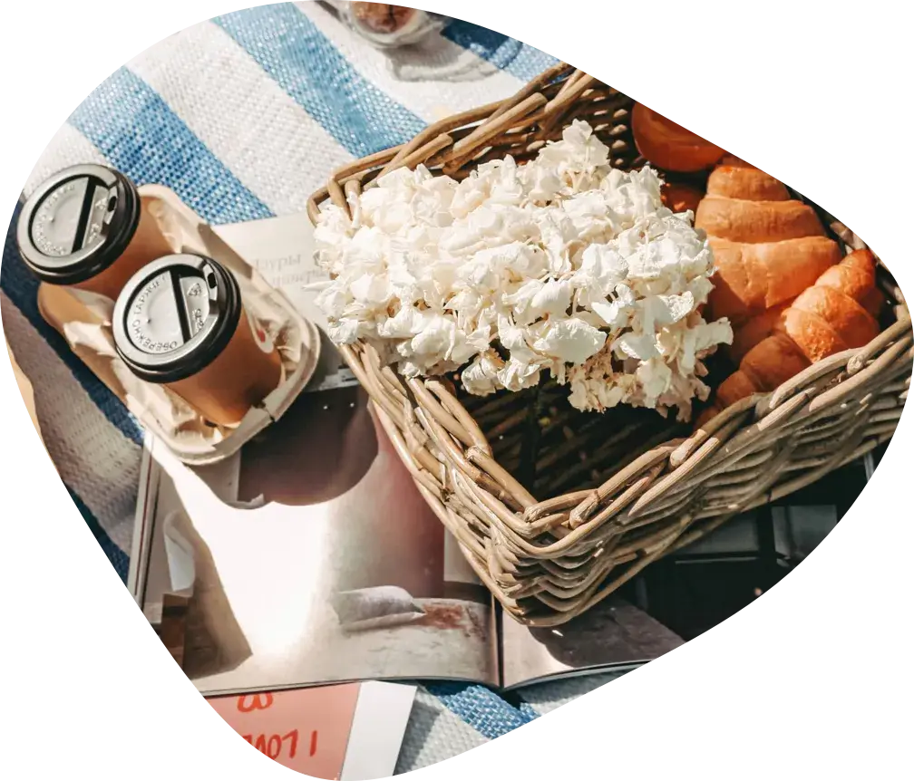 A wicker basket with croissants and white flowers, two takeaway coffees and a magazine on a striped blanket