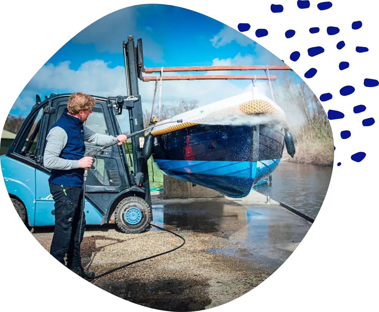 A man pressure-washing a boat that hangs from a forklift