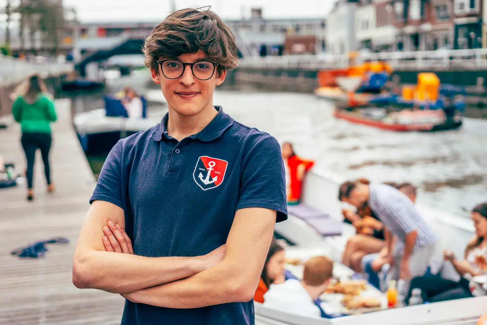A crew member in a navy polo stands on his dock with his arms crossed, guests aboard a boat behind him