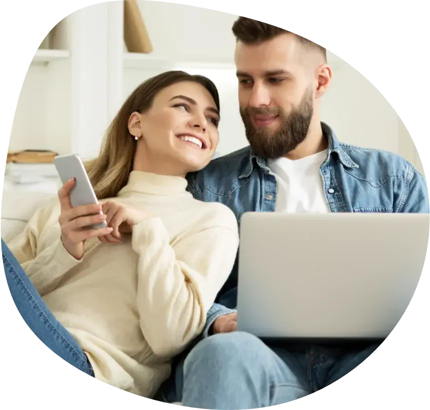 A smiling couple on a sofa, one with a laptop on their lap, the other with a phone