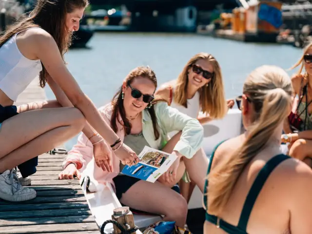 A crew member crouching on the dock shows a leaflet to a group seated in an open boat