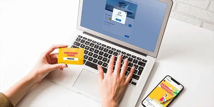 Een hand typt op een laptop, de andere houdt een kaart met een kortingscode vast, met een telefoon ernaast
