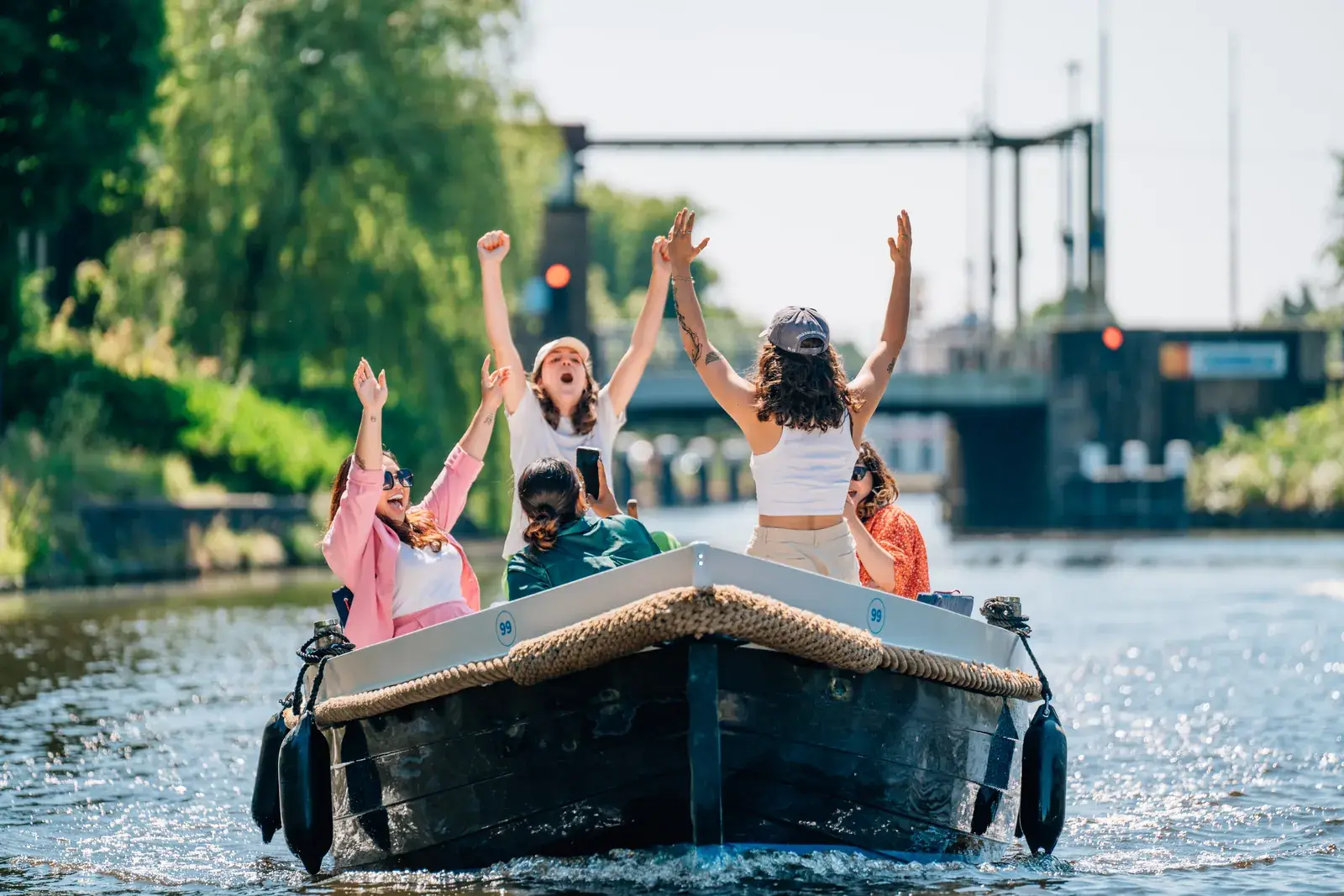 Friends on an open rental motorboat on a sunny canal, three of them cheering with their arms in the air