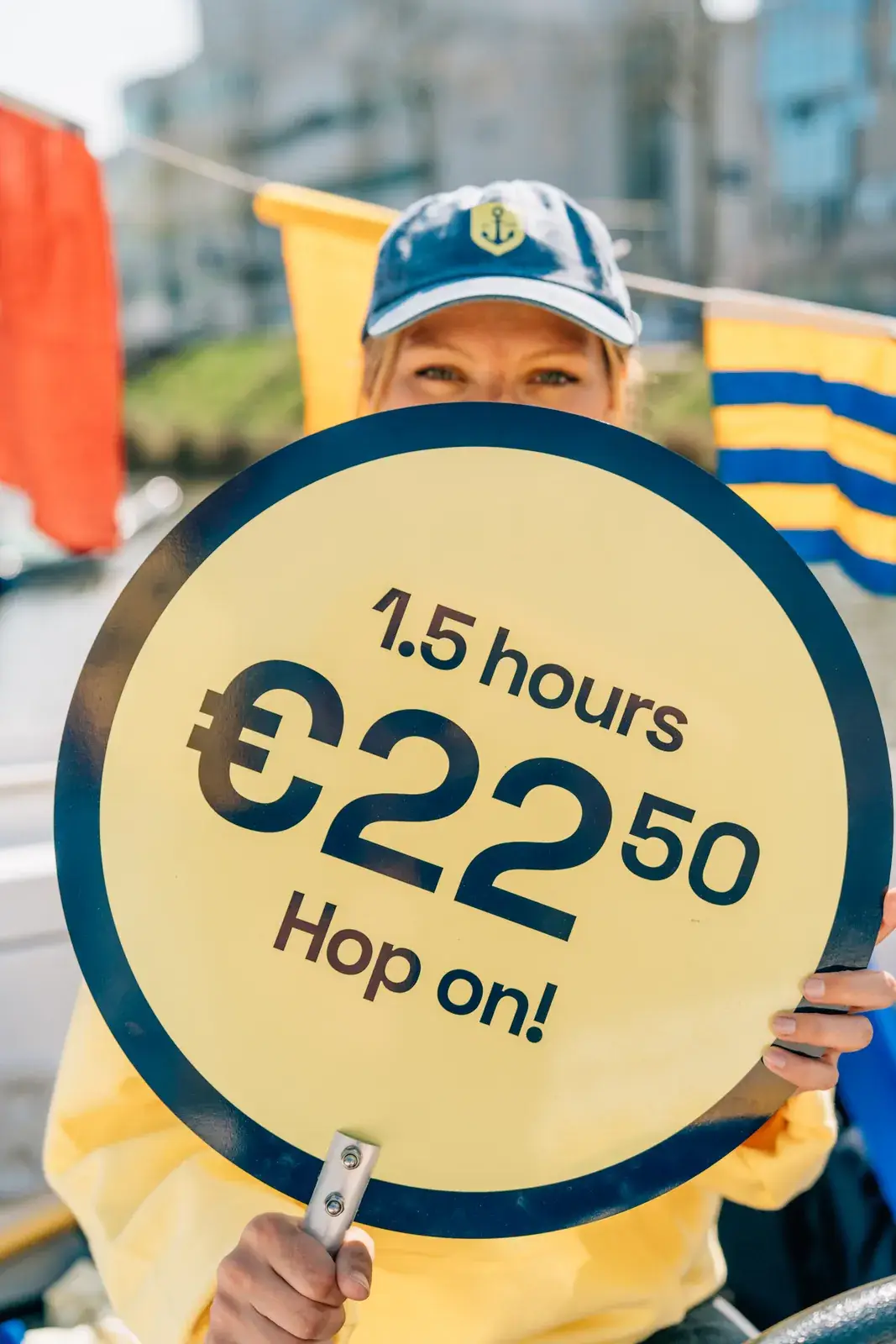 Un membre d'équipage en sweat à capuche jaune qui tient un panneau rond avec le prix d'une croisière sur les canaux
