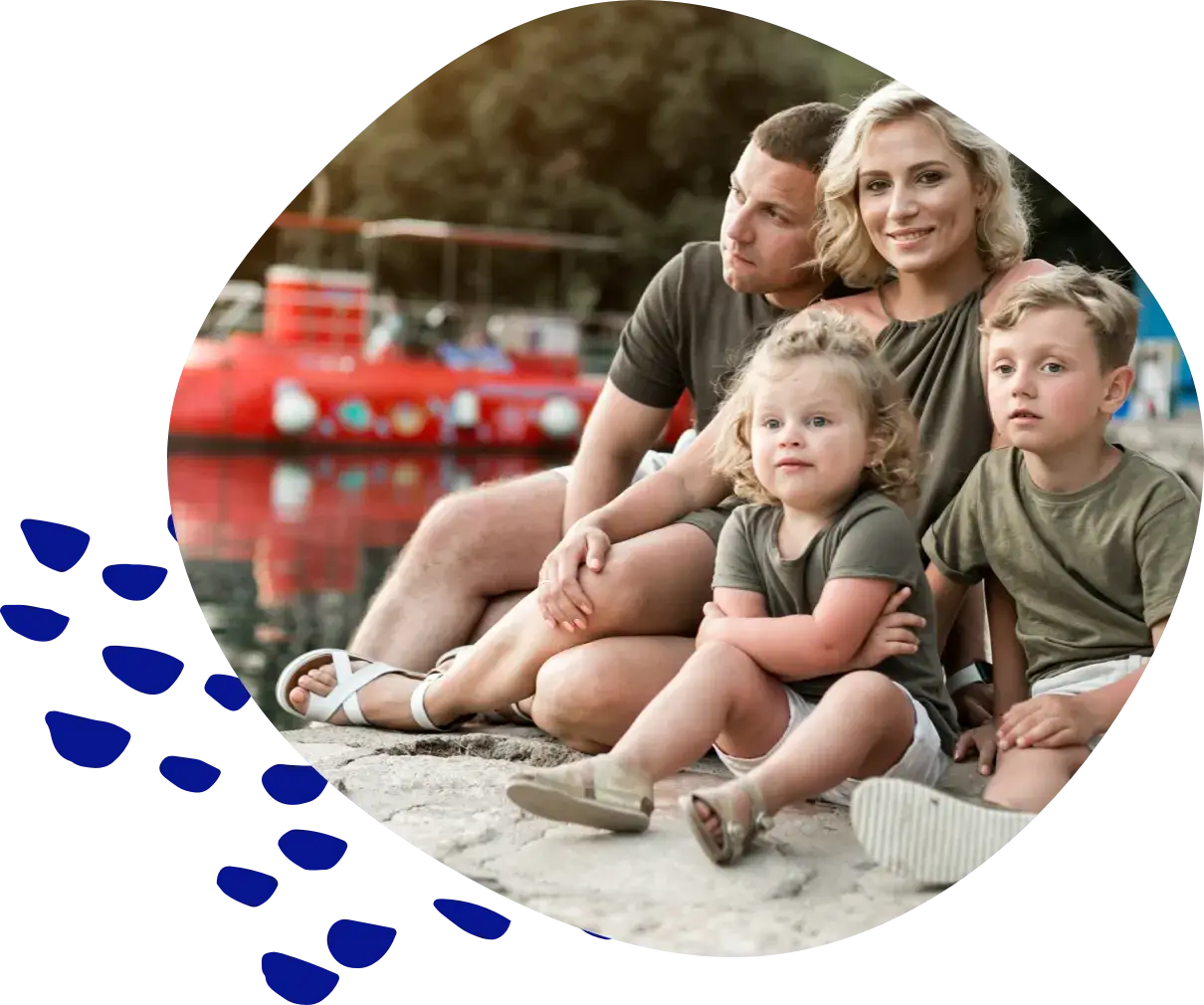 A family with two young children sitting on a stone quay by the water, a red boat moored behind them