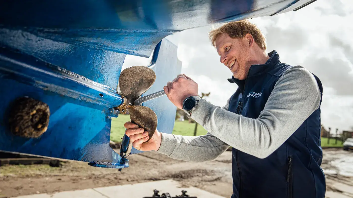 A smiling mechanic works on the propeller under the hull of a boat on land