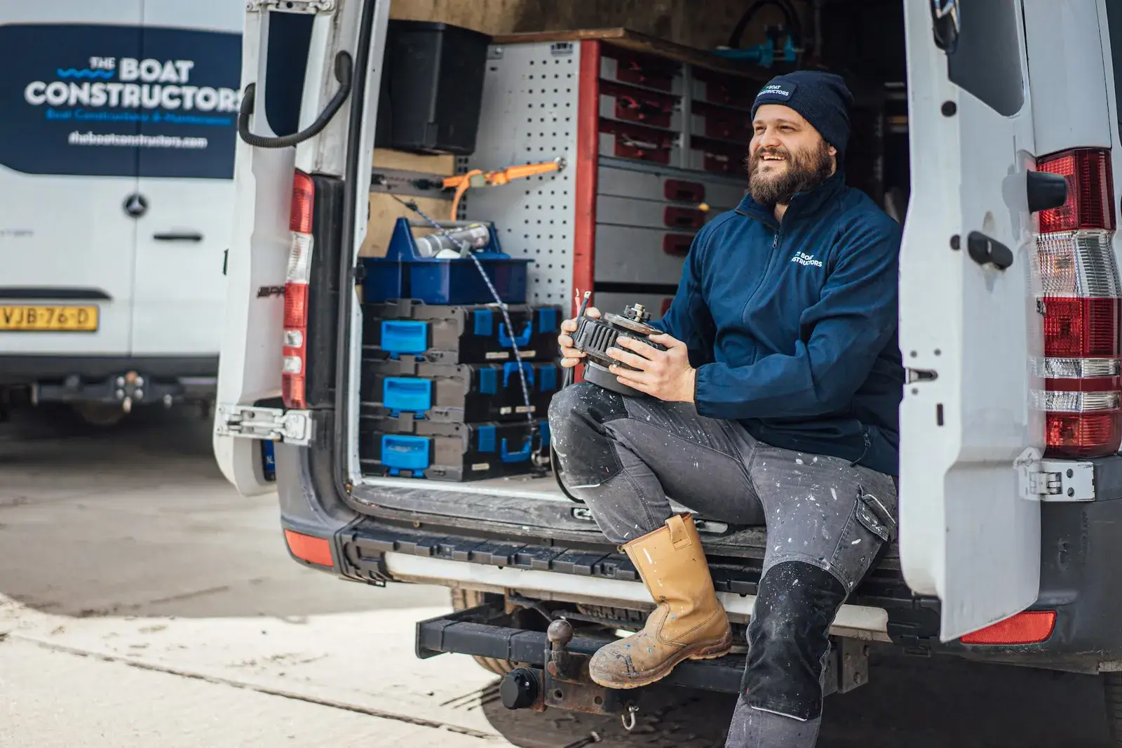 Un mécanicien assis à l'arrière ouvert de sa camionnette d'atelier, pleine de caisses à outils, une pièce dans les mains