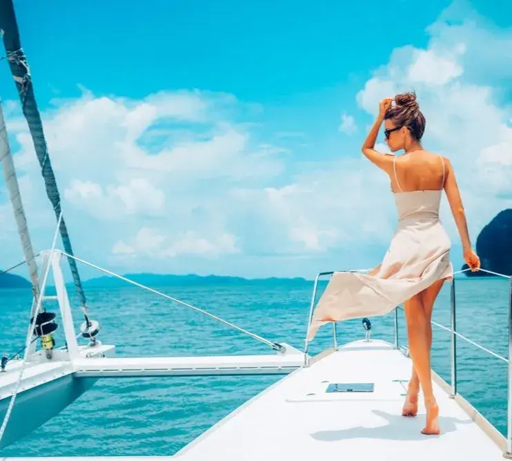 Eine Frau im Sommerkleid steht am Bug eines Katamarans und blickt über das blaue Meer