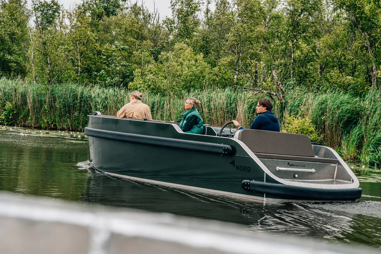 Drei Leute an Bord eines Elektroboots Nord 6 mit Aluminiumrumpf, auf einem Fluss mit Schilf am Ufer