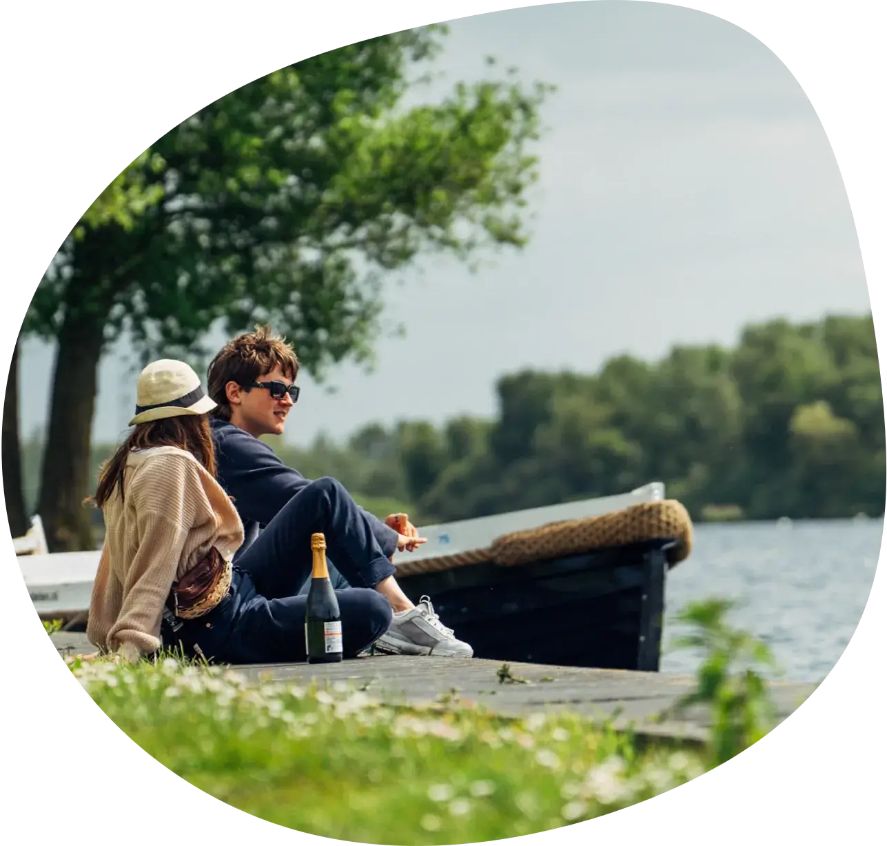 A couple sitting on a dock next to a moored open boat, with a bottle of sparkling wine beside them
