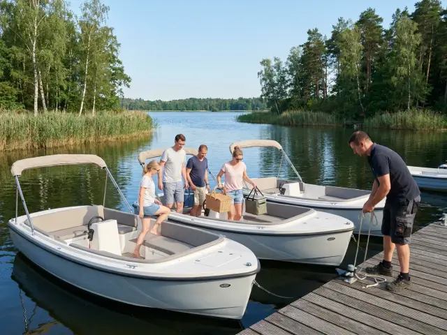 Elektroboote zum Mieten an einer hölzernen Anlegestelle an einem See mit Schilf und Kiefern, eine Gruppe Freunde steigt mit Picknickkorb ein