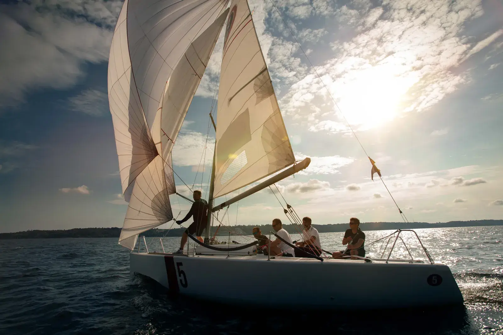 Ein Segelboot mit Crew an Bord unter vollen Segeln, die Sonne tief dahinter