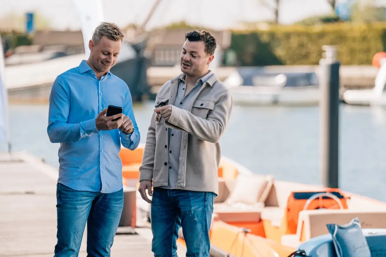 Two men on a dock looking at a phone together, with boats behind them