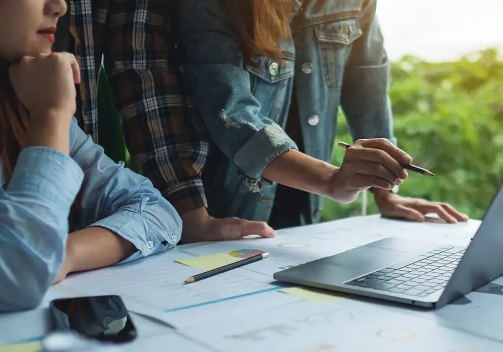 Drie collega's aan een tafel met een laptop en papieren met grafieken, aan het plannen