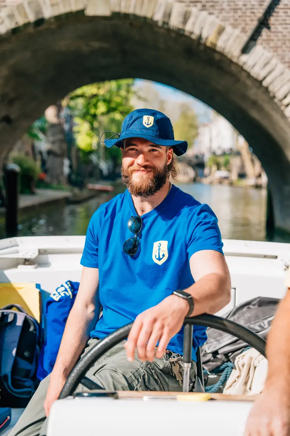 Un skipper souriant en t-shirt bleu et bob à la barre, sous un pont