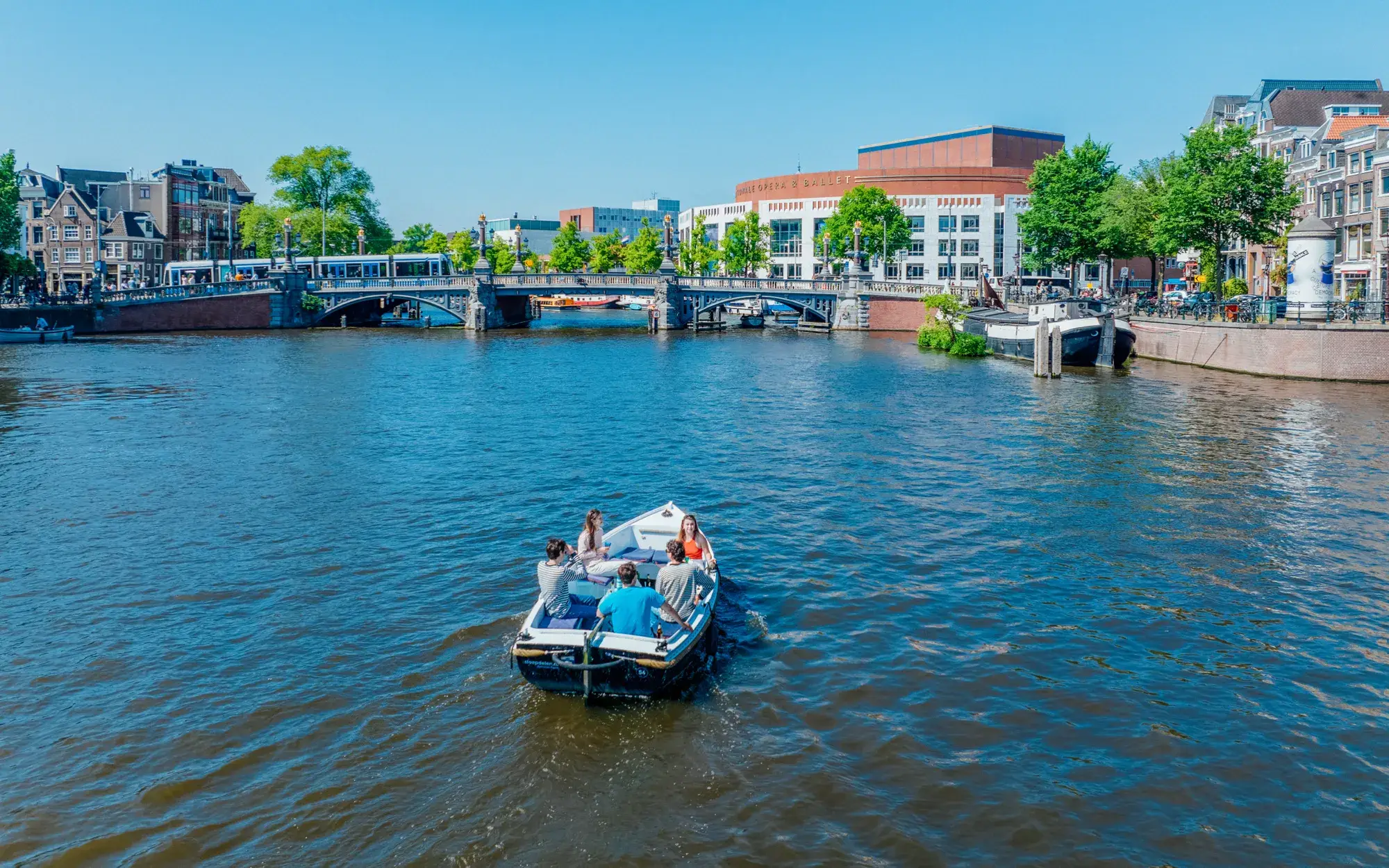 An open motorboat with a group of friends on the wide Amstel in Amsterdam, a bridge and city buildings behind them