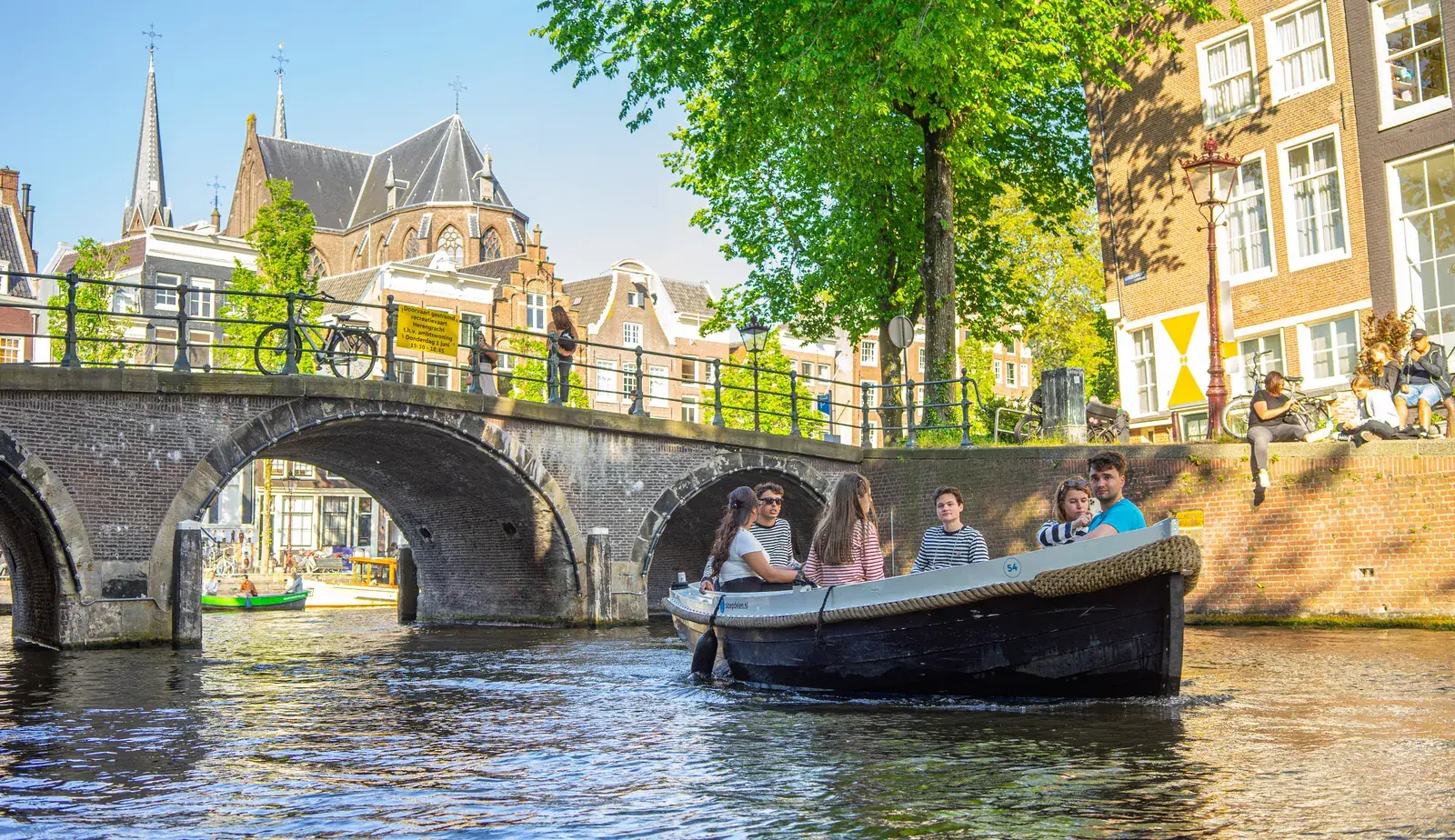 Een sloep met een groep vrienden die onder een bakstenen brug door vaart op een zonnige Amsterdamse gracht