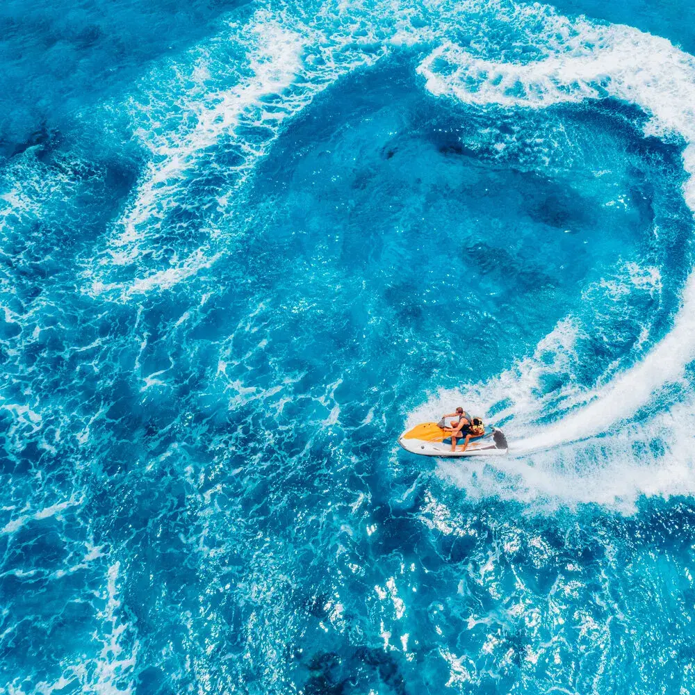 Ein Jetski mit zwei Personen zieht eine Kurve auf hellblauem Wasser, von oben gesehen