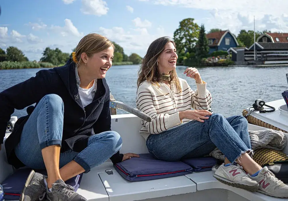 Two women laughing in an open boat on the water, houses on the far bank