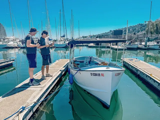 Two of us on a dock in a marina, next to a white rental boat, sailboats behind