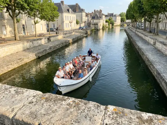 Un bateau-promenade ouvert plein de passagers sur une rivière, entre les quais de pierre d’une vieille ville française