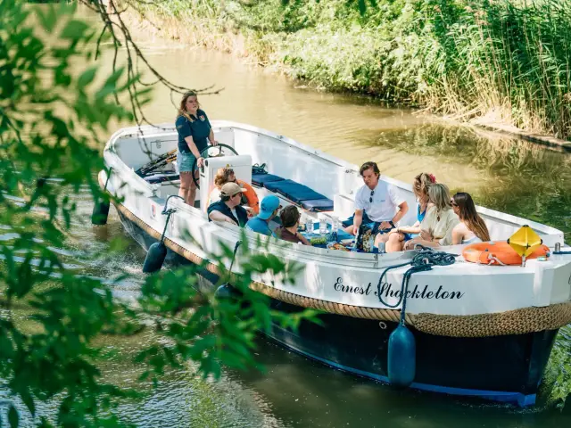 Een open rondvaartboot op een groene rivier, een schipper aan het roer en een groep gasten aan boord, gezien door de bladeren