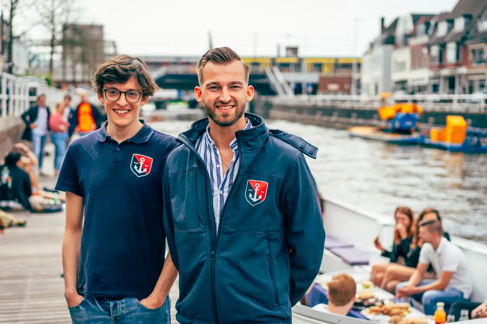 Deux membres de l'équipe d'un loueur de bateaux à Utrecht sourient sur leur quai, avec des clients qui déjeunent à bord derrière eux