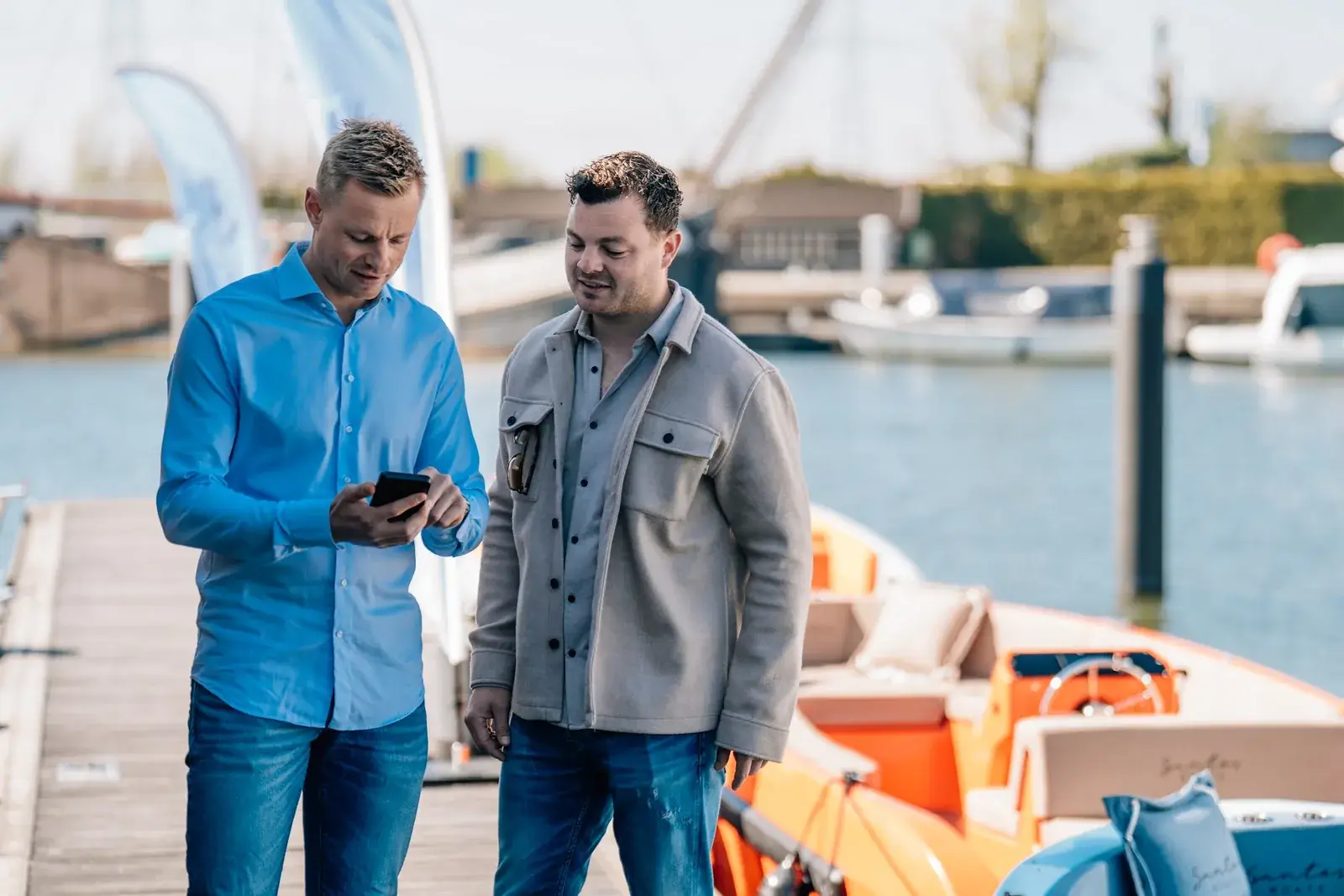 Two men on a pontoon look at a phone together, an orange rental boat moored beside them