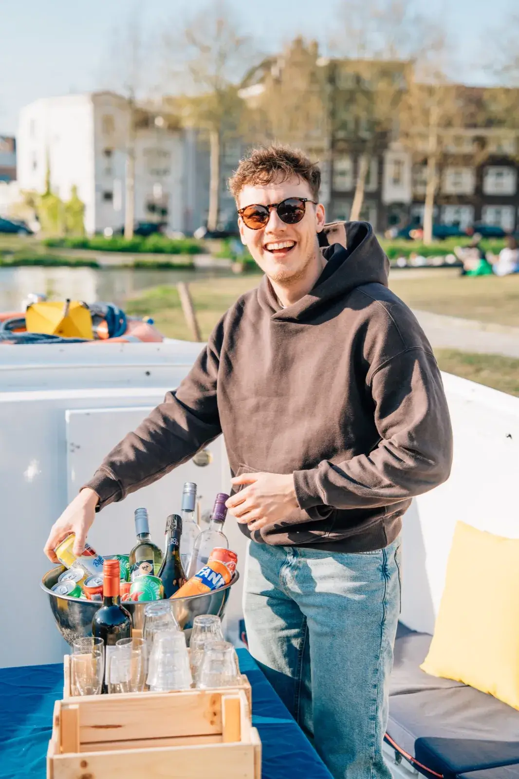 A smiling man takes a can from a bucket of drinks on ice aboard a canal cruise boat in Utrecht, with glasses ready