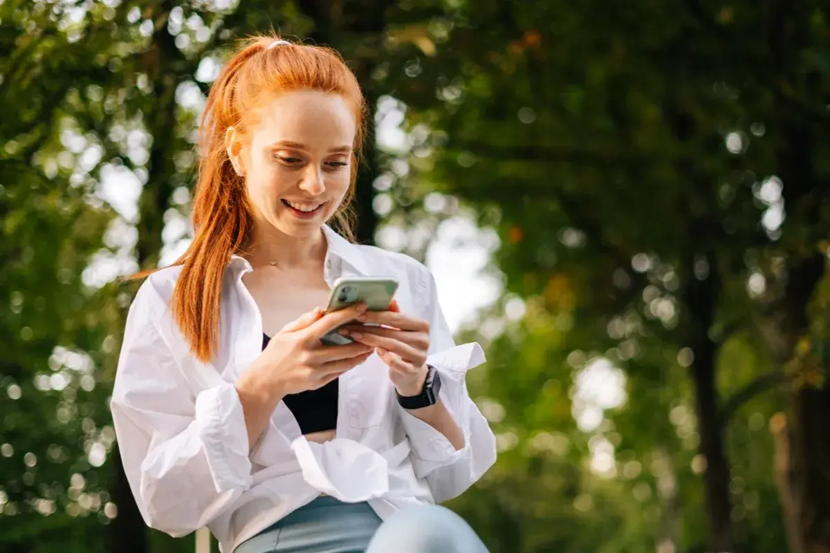 A woman in a white shirt smiles at her phone under the trees, looking something up